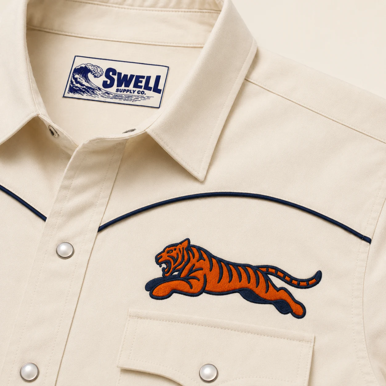 Close-up rendering of the raised tiger embroidery, navy piping, pearl snaps, and Swell neck label.